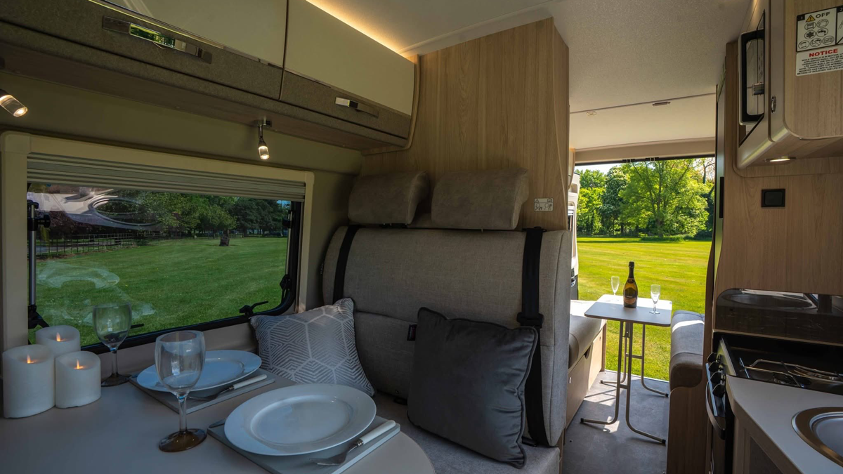 Interior view of a motorhome dining area with set table and large windows looking out onto green countryside
