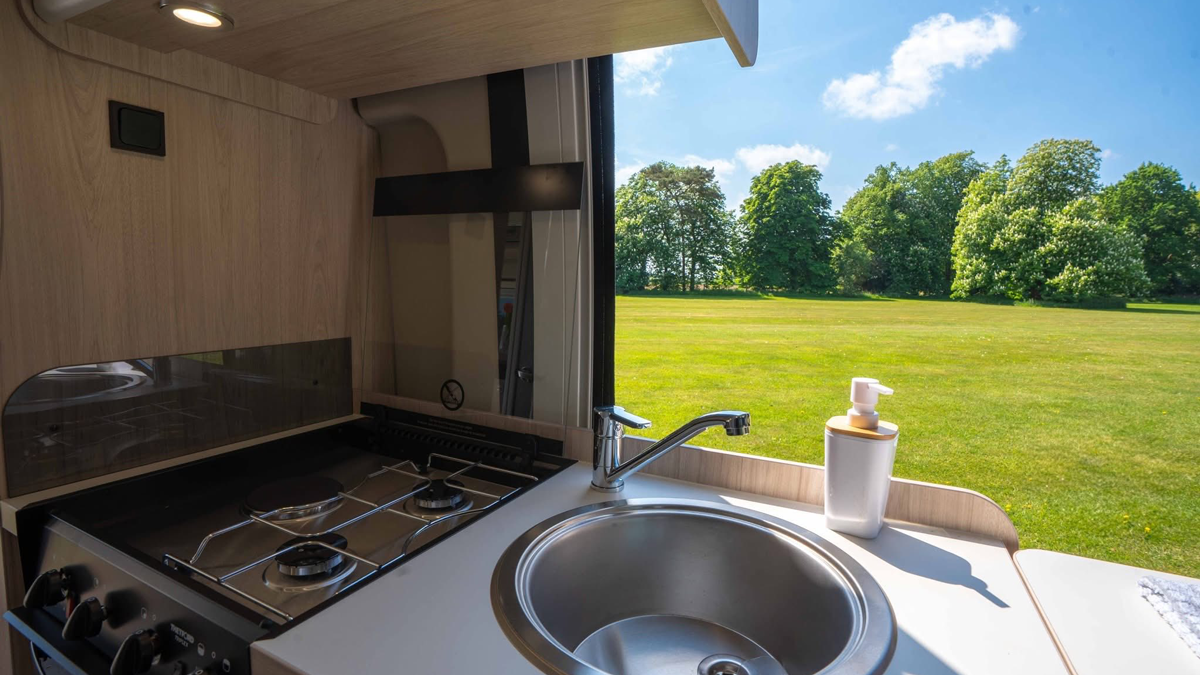 Motorhome kitchen with gas hob, sink, and worktop looking out through the side door onto a sunny green park