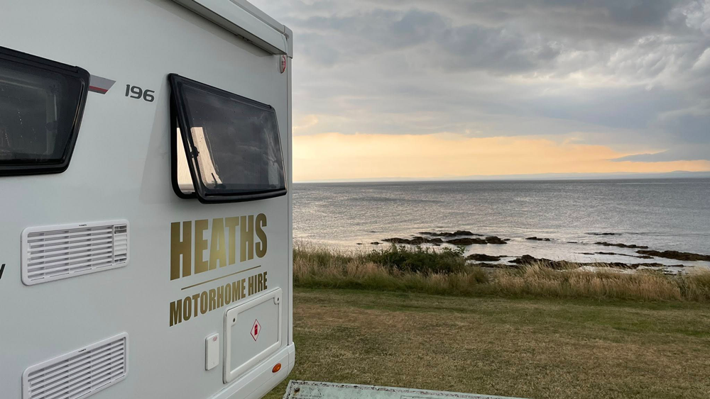 Rear of a Heaths Motorhome Hire motorhome parked at a coastal campsite overlooking the sea at dusk