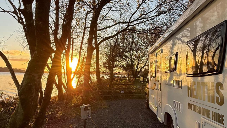 Heaths Motorhome Hire motorhome parked among trees beside a lake at golden hour sunset