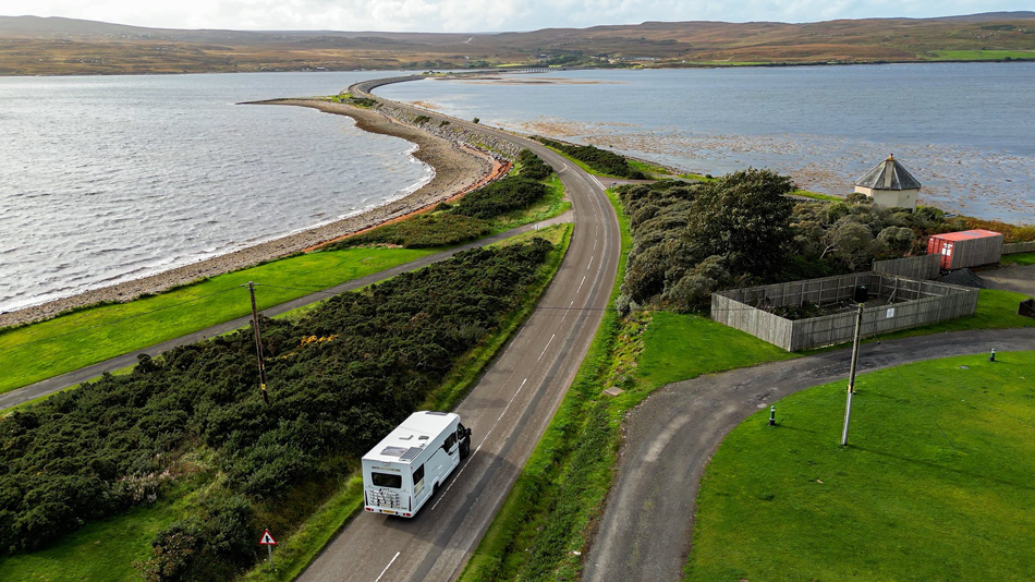 Aerial view of a motorhome driving along a coastal road beside green fields and a bay in Scotland