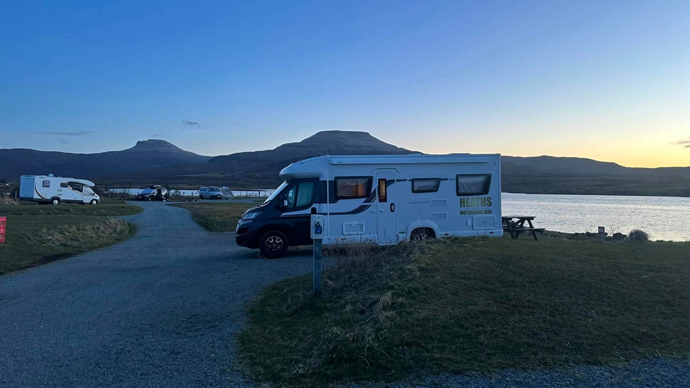 Motorhomes parked at a lochside campsite at dusk with mountains silhouetted against the evening sky